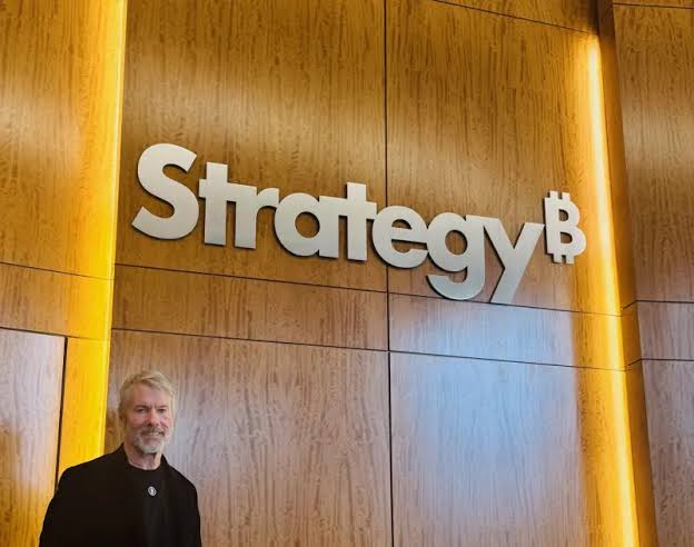 Michael Saylor stands beneath a large "Strategy" logo (formerly MicroStrategy), the company he leads that holds the largest corporate Bitcoin treasury—contextualizing the post's concern that his $225M BTC sale could trigger a major price drop similar to past selloffs.