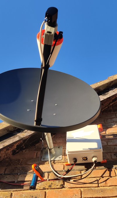 A satellite dish mounted on a brick wall with a white electronic box and battery connected via cables, set against a clear blue sky.