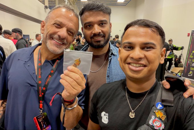 Three men smiling for a photo at what appears to be a convention, with one man holding up a small plastic bag containing a collectible item.