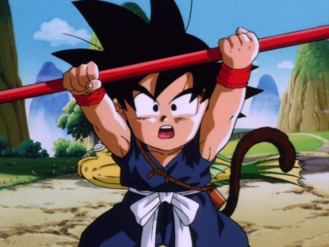 The image features a cartoon character, likely a young boy or a child, wearing a blue outfit with a white bow tie. He is holding a red stick and appears to be playing with it, possibly imitating a dragon or another mythical creature. The background of the image shows mountains in the distance, suggesting that this scene takes place in an outdoor setting. The caption for the image reads "$GOKU".