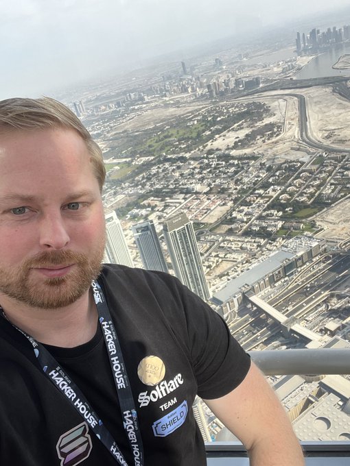 A bearded man wearing a black 'Syscoin' t-shirt and Hacker House lanyard takes a selfie from a high vantage point overlooking a city
