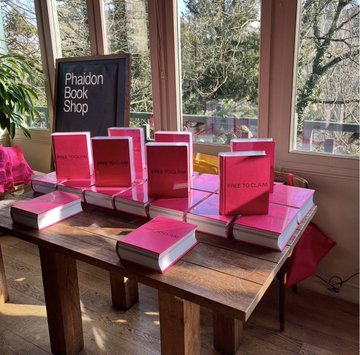 Multiple copies of a bright pink book titled 'Phaidon Book Shop' and 'FRED TOUSSAINT' are stacked on a wooden table near a chalkboard sign reading 'Phaidon Book Shop'.