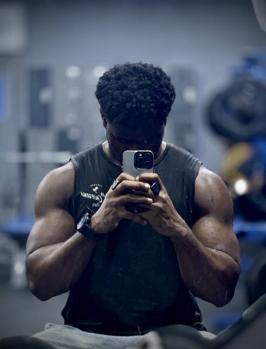A muscular man in a dark sleeveless shirt looks down at his smartphone while sitting in a gym setting.