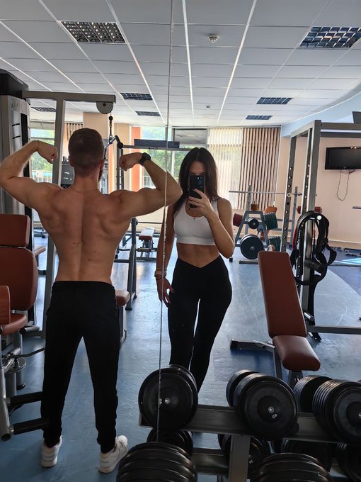 A muscular man and a woman in athletic wear pose for a mirror selfie in a gym, with the man flexing his back muscles and the woman taking the photo with her phone, embodying the post's message about taking action and stopping the cycle of postponing the pursuit of a better, healthier life.