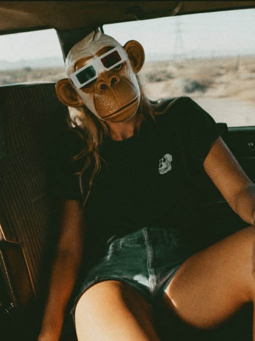 The image features a woman wearing a monkey mask, sitting inside a car with her legs crossed. She has blonde hair and is dressed casually in shorts. The background of the photo shows an open road or highway, suggesting that they might be on their way somewhere. The caption at the bottom of the image reads "GM where are we headed? bored ape yacht club". This phrase could be interpreted as a reference to the monkey mask worn by the woman and her surroundings, possibly indicating that she is in a themed location or event related to monkeys or apes.