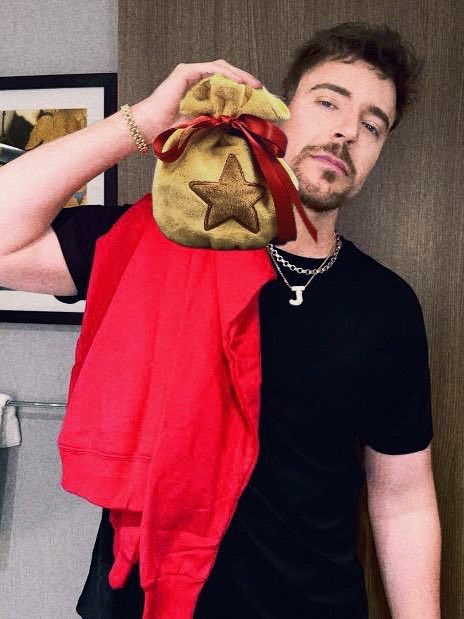 A man with a goatee, wearing a black t-shirt and a silver chain necklace with a 'J' pendant, holds up a red hooded garment with a gold star bag over his shoulder.