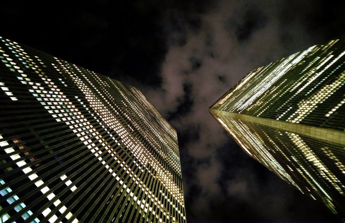 Two illuminated skyscrapers tower against a dark, cloudy night sky, viewed from a dramatic low angle that emphasizes their towering height and the grid patterns of their lit windows.