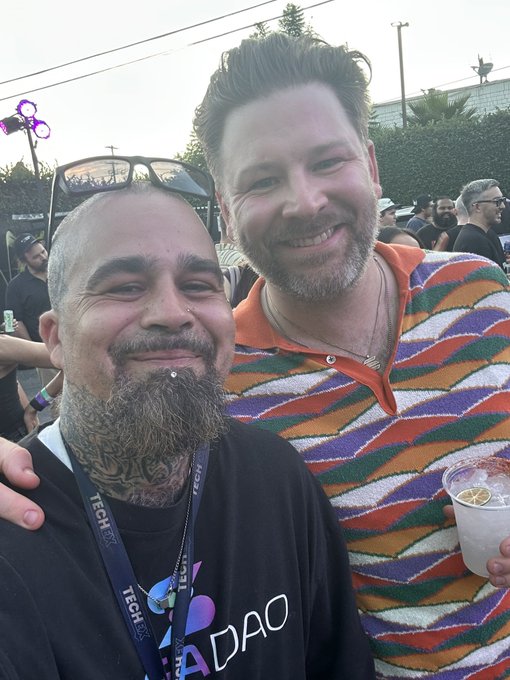 Two smiling men posing together outdoors at a tech event; one wears a lanyard and a black 'ADAO' shirt, while the other wears a colorful striped polo.