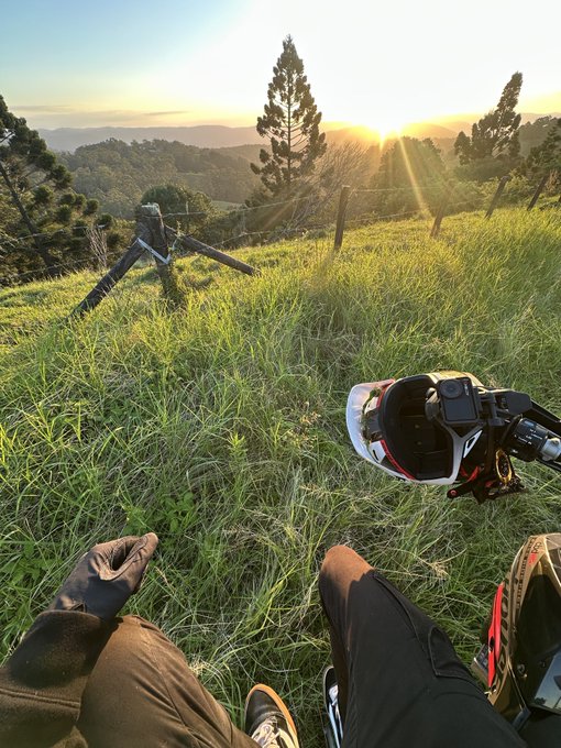 A first-person view of someone sitting on a grassy hillside at sunset, with a motorcycle parked nearby and a scenic valley visible in the distance.
