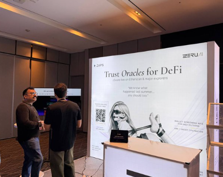 Two men in casual clothing stand in a conference area near a booth with signage reading 'ZERU AI' and 'Trust Oracles for DeFi', featuring an anime-style character illustration.