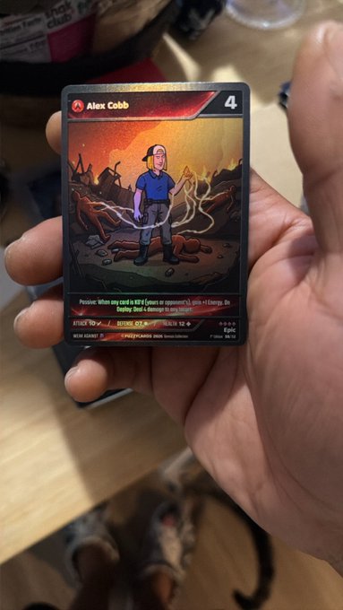 A hand holds up an "Alex Cobb" character card from the $FUZZY TCG trading card game, with a Defend ×1 ability, while a tired-looking calico cat rests on the floor below—tying into the post celebrating Cobb's inclusion in the FUZZY TCG as an actual collectible card.