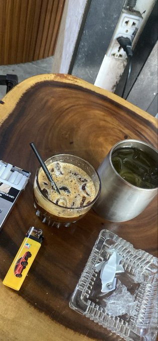 A wooden tray holds an iced coffee drink, a metal cup of green tea, a yellow Ferrari-branded lighter, a pack of cigarettes, and an ashtray with a used cigar.