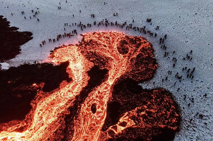 An aerial view shows an active volcanic eruption with glowing orange-red lava flowing through a dark, solidified crust, while small figures of onlookers stand on the surrounding gray terrain.