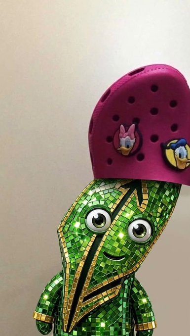 A green mosaic-covered figurine wearing a pink Croc shoe on its head with Daisy and Donald Duck charms attached.