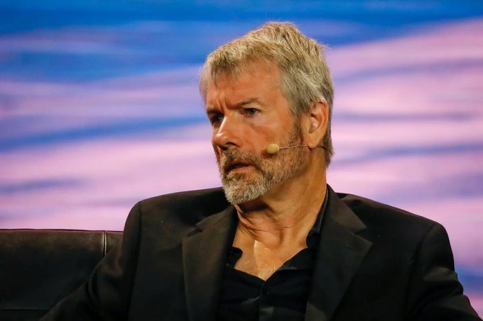 A gray-haired man with a beard and a headset microphone, wearing a black suit jacket over a dark shirt, sits on stage against a blue and purple backdrop.