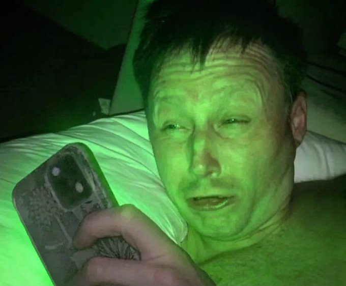 A close-up photo of a man's face illuminated by green light as he lies in bed looking at a smartphone.