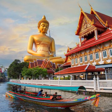 A large golden Buddha statue sits next to a traditional Thai temple with ornate red and gold roofs, while a colorful long-tail boat with passengers floats on the water in the foreground.