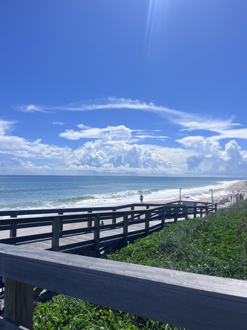 A scenic view from a wooden boardwalk looking out toward a beach and ocean under a bright blue sky filled with fluffy white clouds.