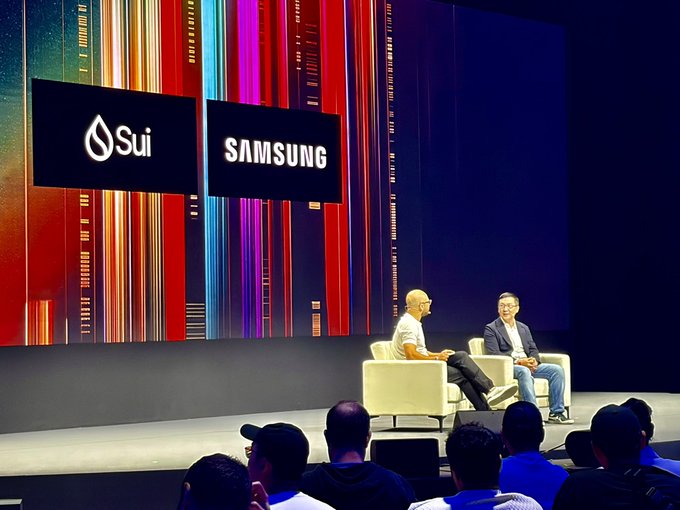 Two men sit in armchairs on a stage during a panel discussion, with large screens behind them displaying the 'Sui' and 'SAMSUNG' logos.