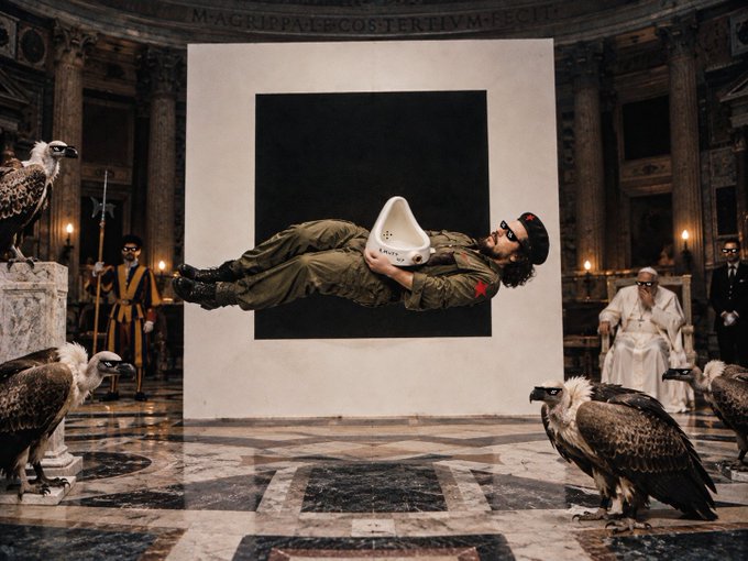 Artistic photograph of a person floating horizontally in front of a black square canvas inside an ornate building, surrounded by vultures and onlookers, with the inscription 'M·AGRIPPA·L·F·COS·TERTIVM·FECIT' on the wall.