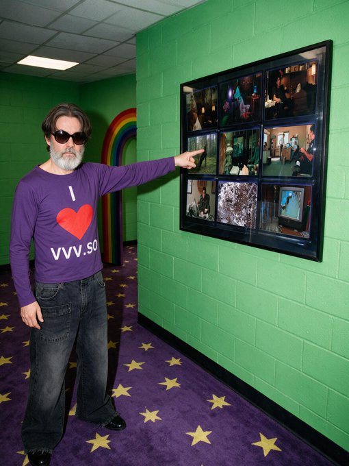 A bearded man wearing sunglasses and a purple "I ❤️ www.something" shirt points at a wall-mounted frame displaying a grid of nine photographs in a green-walled room with a star-patterned purple carpet. The colorful, eclectic setting with a rainbow arch and whimsical decor complements the post's discussion of curated art collections and traits.