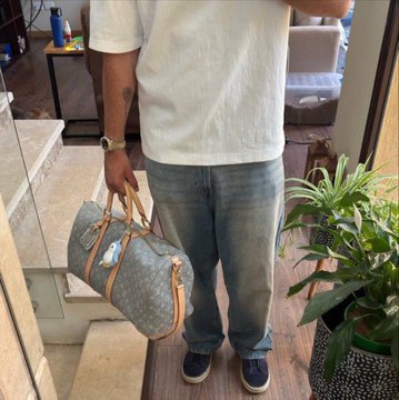A person wearing a white t-shirt and jeans stands holding a Louis Vuitton Speedy bag containing what appears to be an American Express card, photographed in a mirror selfie. The image humorously illustrates the "Crypto bros 2025 vs now" meme by showing someone who supposedly struck it rich in crypto now quietly flexing modest luxury goods instead of flashy displays.