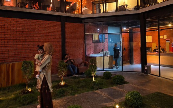 A woman wearing a beige hijab holds a child while standing outside an evening-lit building with exposed red brick walls and a small landscaped garden.
