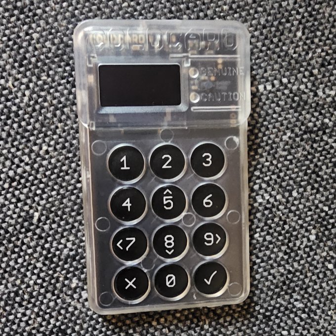 A transparent hardware cryptocurrency wallet with a small screen and numeric keypad (1-9, 0, along with cancel/confirm buttons) labeled "GENUINE" and "CAUTION" rests on a gray carpet. The visible "GENUINE" and "CAUTION" indicators, combined with the casual "Damn" caption, suggest the user has likely confirmed the device as authentic but is wary about using it—perhaps due to concerns about its security or condition.
