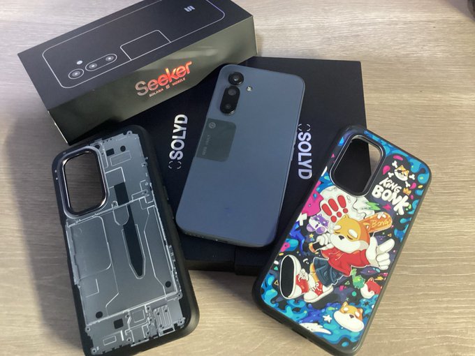 A photograph showing a black smartphone with its packaging box and two cases laid out on a wooden surface: a translucent gray case and a colorful one featuring Mario.