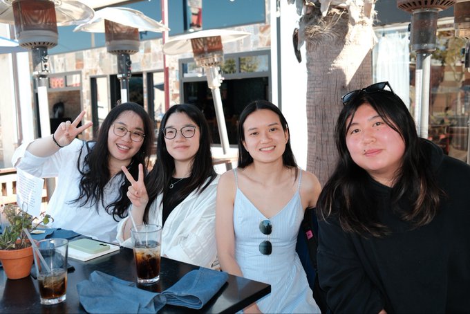 Four smiling young women sitting together at an outdoor restaurant table with drinks, posing for a photo with two flashing peace signs.