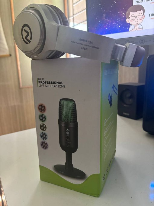 A pair of silver Raijintek NYX headphones rests on top of its product packaging for an 'RGB PROFESSIONAL LIVE MICROPHONE', sitting on a desk with a monitor in the background.