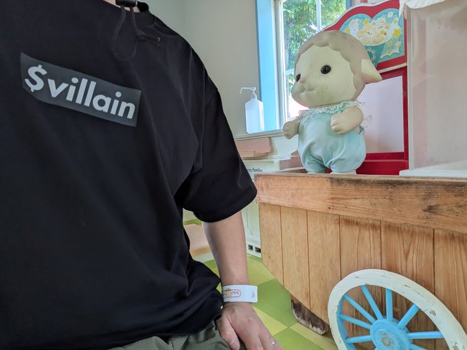 A person wearing a black t-shirt with a '$villain' patch sits near a wooden cart that holds a plush lamb toy in a light blue outfit.