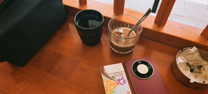 A wooden table holds a black bag, a dark ceramic cup, a glass with iced coffee and a spoon, an 'ESSE' cigarette pack, a smartphone with a circular camera, and a round ashtray.