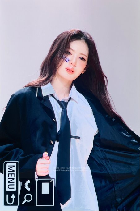 A young woman with long dark hair poses against a white backdrop, wearing a white collared shirt, black tie, and oversized black jacket draped over her shoulders. The image has a retro camera interface overlay with "MENU" and a shutter button visible, and the watermark reads "260823 #KiiiKiii #키키 #SUI #수이 #スイ @We_KiiiKiii," identifying the subject as SUI from the K-pop group KiiiKiii in a stylized photoshoot.