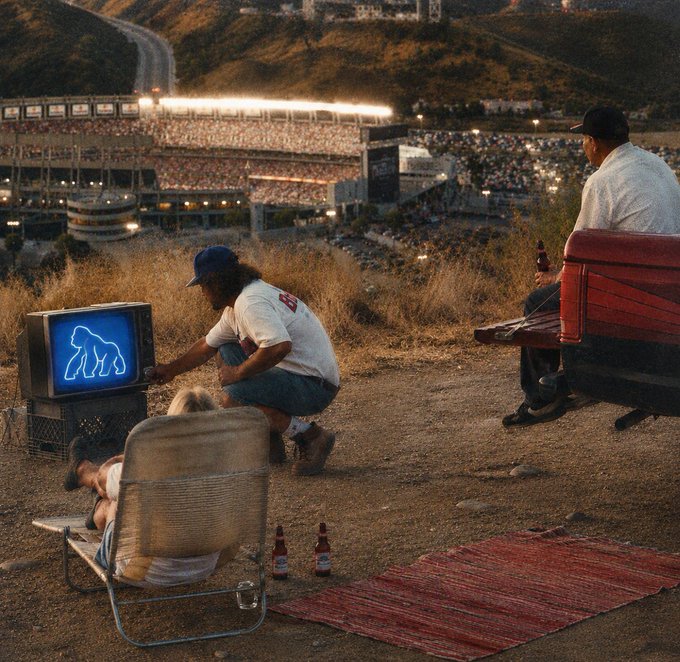 A group of friends gather on a hilltop at dusk, watching a vintage TV displaying a gorilla silhouette while overlooking a stadium lit up below. The scene's nostalgic, laid-back vibe of casual viewing ties in with the post's message to tune in and follow the curated APECHAIN list for an upgraded algorithm.