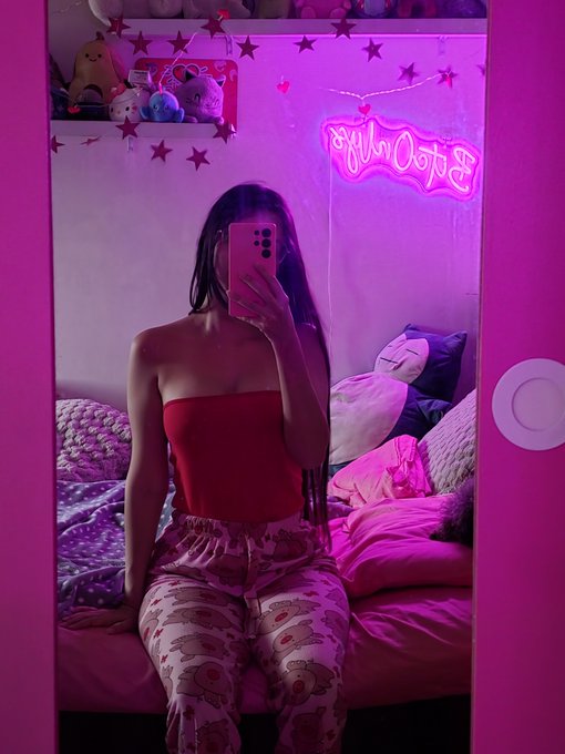 A mirror selfie of a woman in a red top and printed pants, bathed in pink neon with a sign reading 'OK, that's me' visible in the reflection.