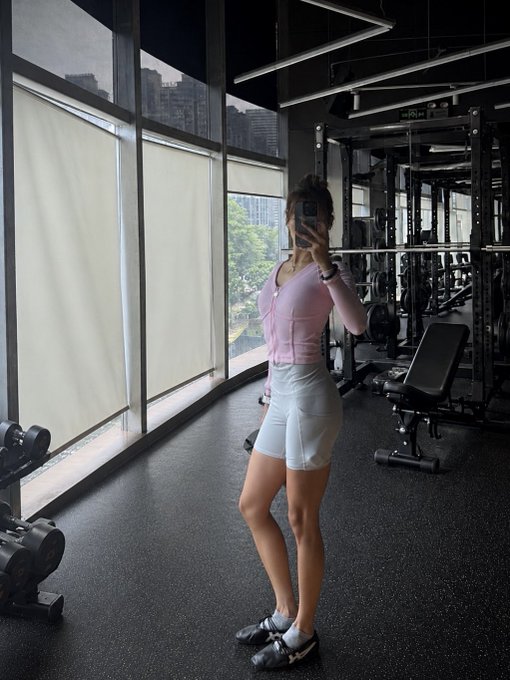 A woman in a pink cardigan and white shorts takes a mirror selfie in a gym with large windows overlooking a city, holding a phone that obscures her face.