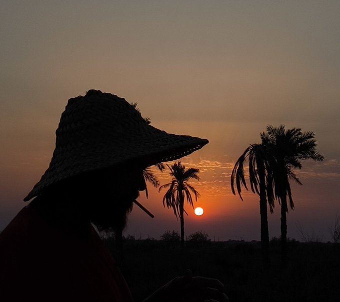 Silhouette of a person wearing a woven hat and smoking against a warm sunset with palm trees visible in the background.