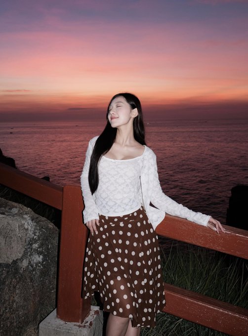 A woman in a white patterned top and brown polka-dot skirt stands by a wooden railing, smiling peacefully with her eyes closed against a sunset seascape.