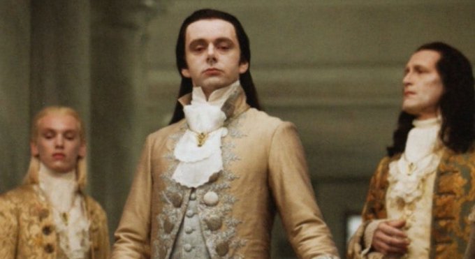 A pale man with long black hair wearing an ornate gold and white period costume stands between two similarly dressed figures in a palatial setting.