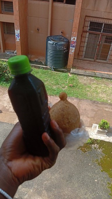 A hand holds a bottle of dark liquid and a wrapped piece of food, standing outside near a black water tank labeled "9000 litres" in front of a brown building, suggesting the everyday items and surroundings trigger memories of the person being remembered.