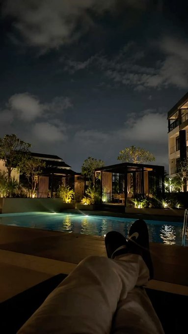 A first-person perspective photo taken at night, showing the photographer's feet wearing slippers resting by the edge of a softly lit swimming pool, with illuminated resort buildings and trees in the background.