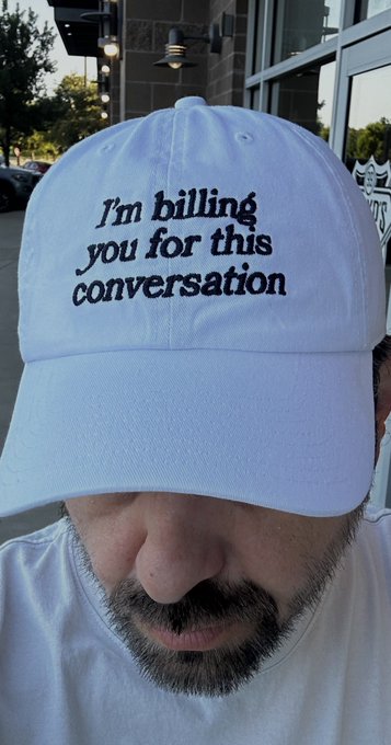 A man wearing a white baseball cap with the embroidered text "I'm billing you for this conversation" in black gothic-style font, with only his nose, mustache, and goatee visible below.