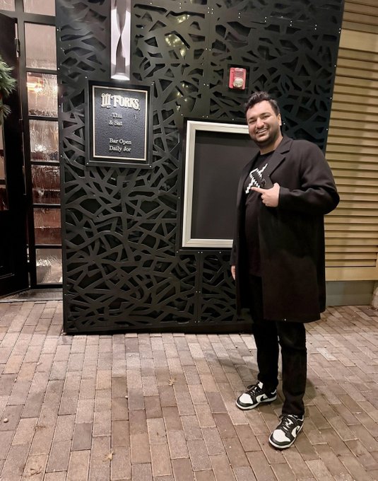 A smiling man wearing a black coat over a black graphic t-shirt and black and white sneakers poses outside '10 forks' restaurant, which displays 'Daily Open / Daily Bar' on its chalkboard sign.