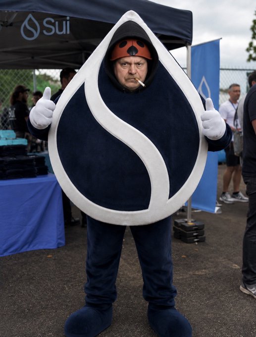 A man in a Sui-themed costume—featuring the iconic dark blue water droplet logo with white outline, a red helmet, and white gloves—stands at the "Sui" branded booth giving two thumbs up while smoking a cigarette, embodying the blockchain's playful branding at the Sui Basecamp event.
