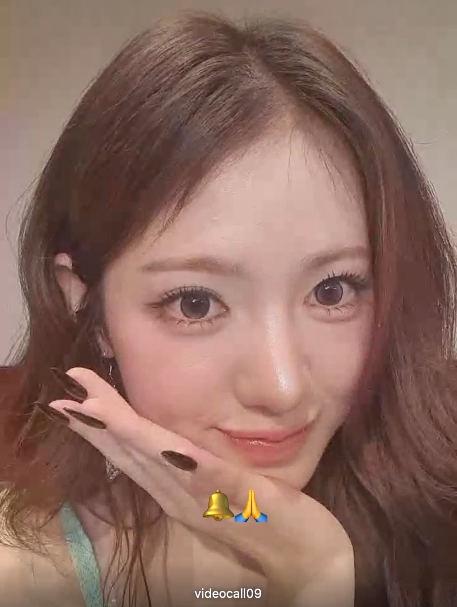 A close-up selfie of a woman with long brown hair resting her chin on her hand; bell and prayer hand emojis are overlaid with the text 'videocall09'.