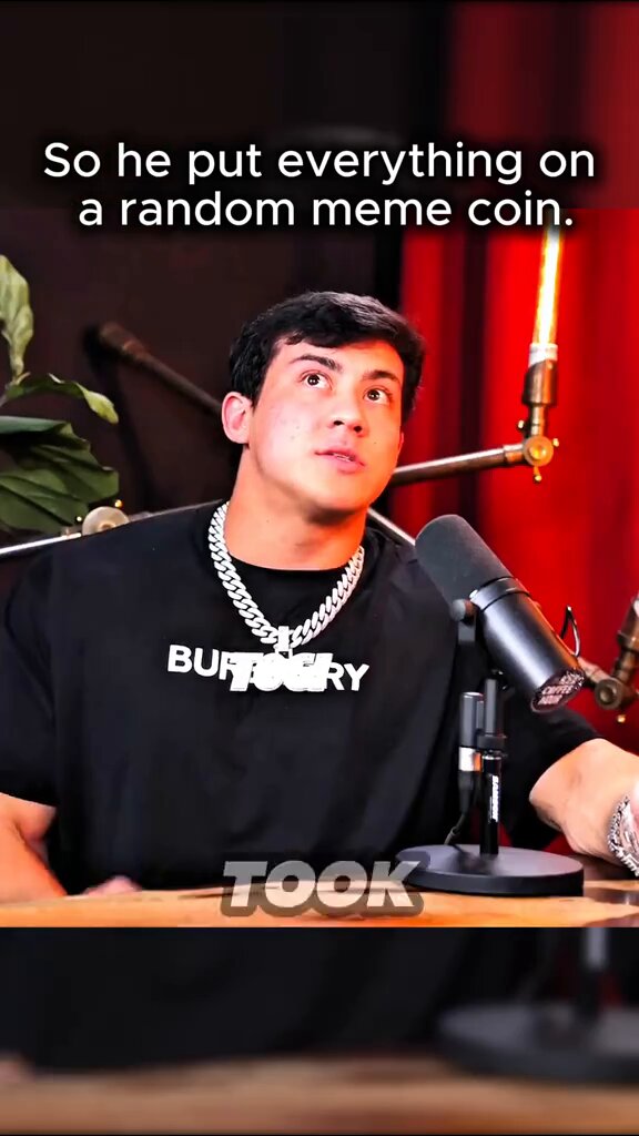 In the image, there is a man sitting at a table wearing a black shirt with white writing. The background shows an empty stage or studio set up with microphone equipment placed in front of him. There are also two other microphones visible in the scene. Above the microphones, there's text on display that reads "so he put everything on a random meme coin".