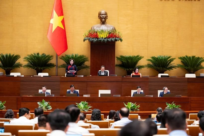 An official assembly hall with the Vietnamese flag, featuring a bust of Ho Chi Minh and officials seated at a long table facing an audience.