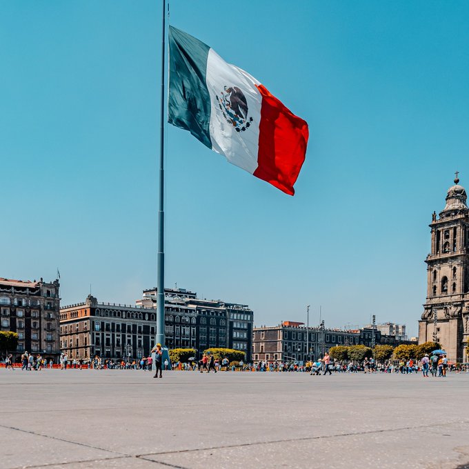 A giant Mexican flag flies at half-mast in Mexico City's Zócalo, with the Metropolitan Cathedral visible in the background. The image appears to be a generic stock photo of the Mexican flag rather than directly related to the news about authorities seizing illegal crypto mining rigs in Tlaola, Puebla.