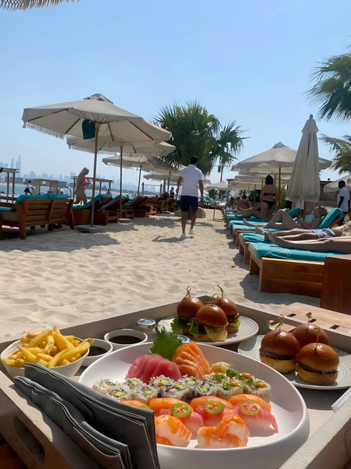A luxurious beachfront spread featuring sushi, sliders, and fries on a sandy shore with palm trees and loungers, capturing the perfect "good morning" beachside brunch vibe.